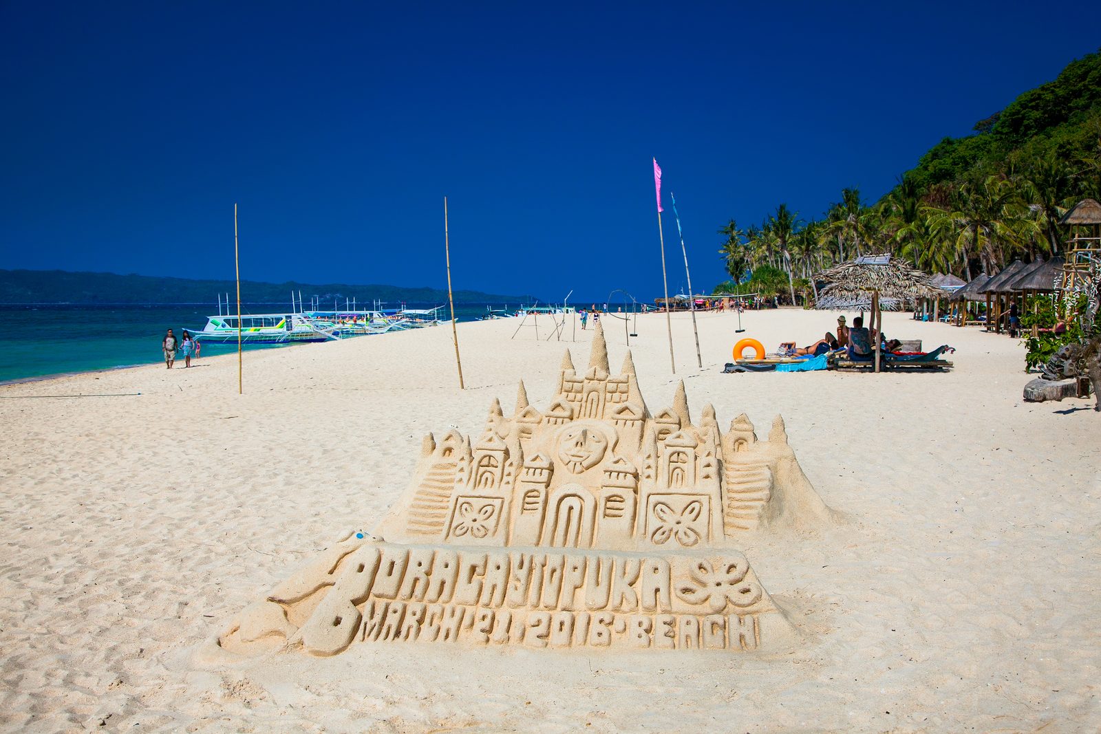 Puka Beach on a bright day, bangkas moored down the shore and a carved sandcastle on the sand.