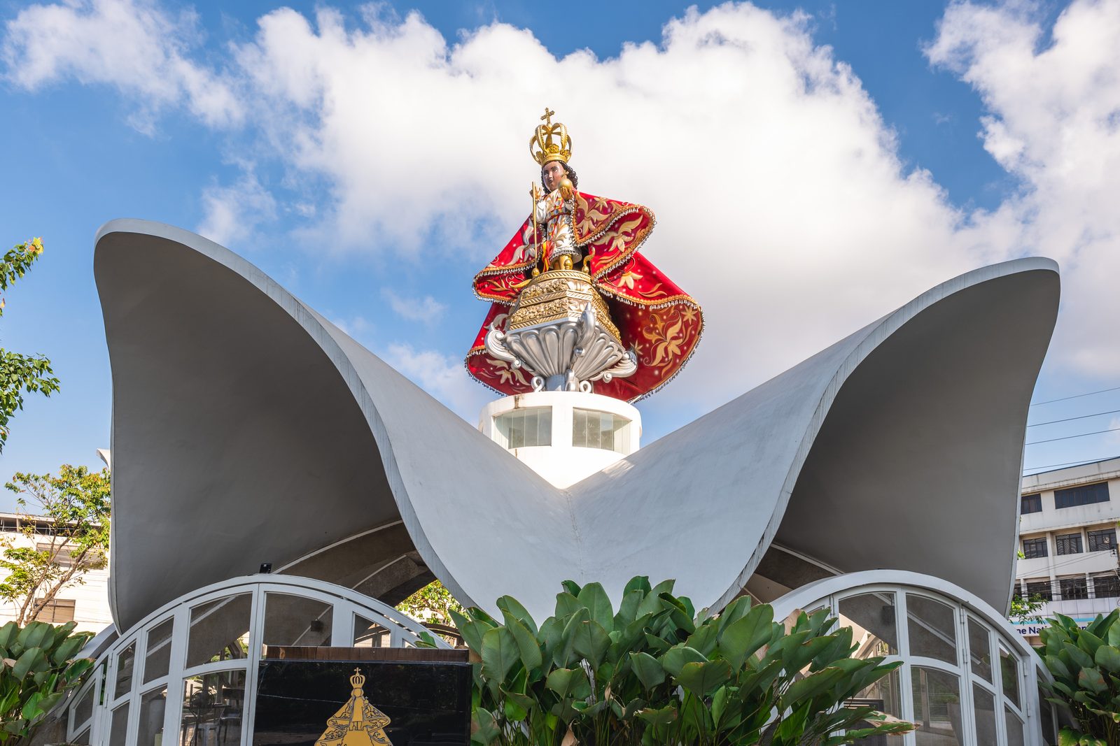 An image of the Santo Ni&ntilde;o in red robes above the white sculptural candle shrine outside the basilica.