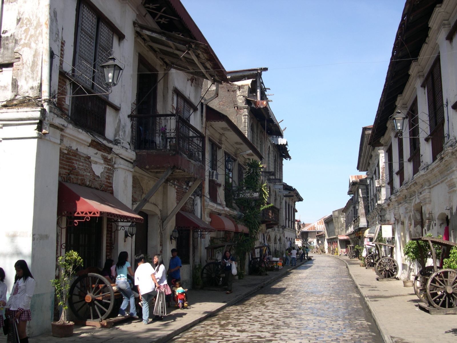 Calle Crisologo&rsquo;s cobbles and ancestral houses in daylight, walkers in the shade of the eaves.