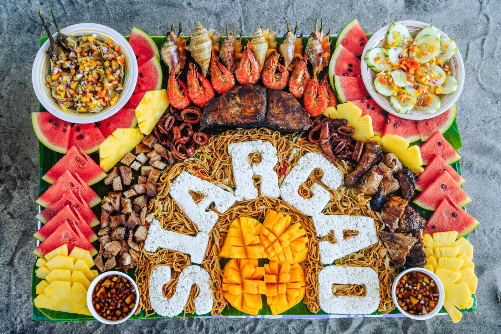 A boodle spread on banana leaves spelling Siargao in noodles, ringed by grilled seafood and fruit.