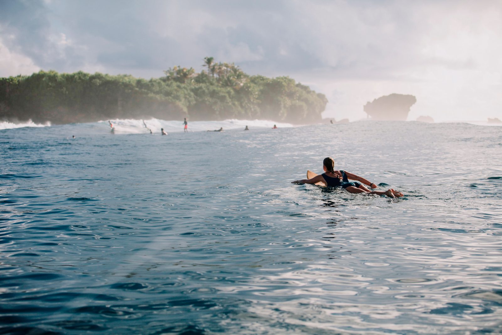 A surfer paddling out toward the break, the island&rsquo;s palm line behind.