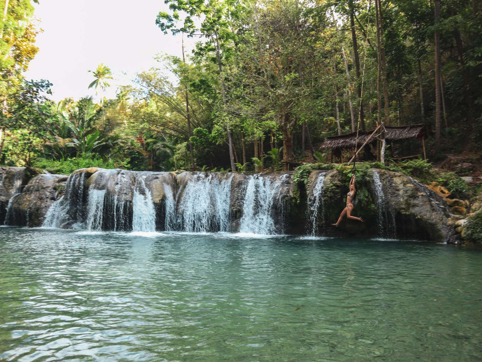 Cambugahay Falls: a swimmer mid-air over the green pool, tiers of spring water behind.