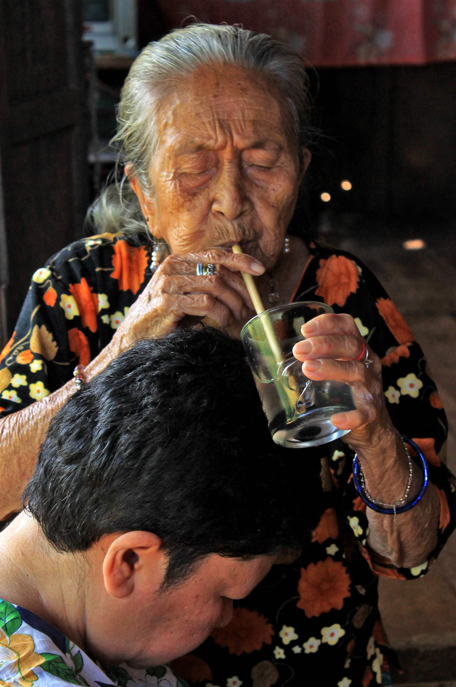 A Bolo Bolo healer blowing through a bamboo straw into a glass of water over a bowed visitor.